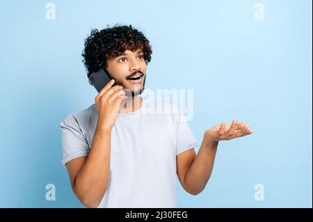 Gesprächskonzept für Mobiltelefone. Positiver arabischer oder indischer attraktiver Kerl, der sein Handy benutzt, auf dem Smartphone spricht, auf einem isolierten blauen Hintergrund steht, wegschaut und freundlich lächelt Stockfoto