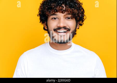 Nahaufnahme eines hübschen attraktiven, lockigen indianers oder arabers, der ein weißes Basic-T-Shirt trägt, auf einem isolierten orangefarbenen Hintergrund steht, die Kamera anschaut und freundlich lächelt Stockfoto