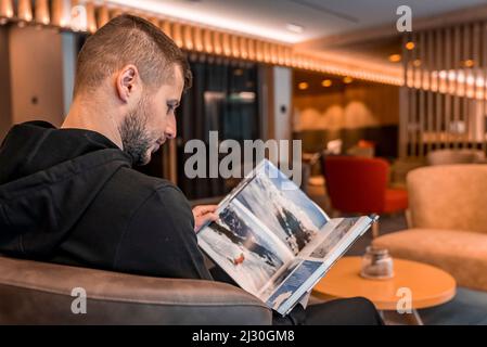 Mann liest den Reisekatalog, während er in der Hotellobby auf einem Sessel sitzt Stockfoto