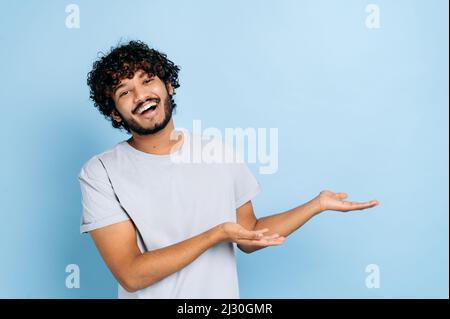 Freundlicher, hübscher, lockiger inder oder araber im lässigen T-Shirt, schaut auf die Kamera und zeigt die Hände zur Seite, auf einen leeren Raum, steht auf einem isolierten blauen Hintergrund und lächelt. Copy-Space-Konzept Stockfoto