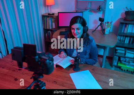Schöne hispanische Frau vor einer Videokamera, die in ihrem Studio einen Blog mit roten und blauen Lichtern in ihrem Haus aufzeichnet. Übertragung Ihres Kommens Stockfoto