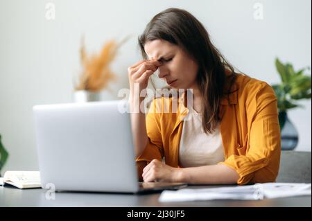 Kopfschmerzen und Migräne. Erschöpfte kaukasische junge Erwachsene Frau, Top-Manager, Büroangestellte, müde von der Arbeit, gestresst, Machte eine Pause von der Arbeit, braucht Ruhe, massiert den Nasenrücken, schließt die Augen Stockfoto