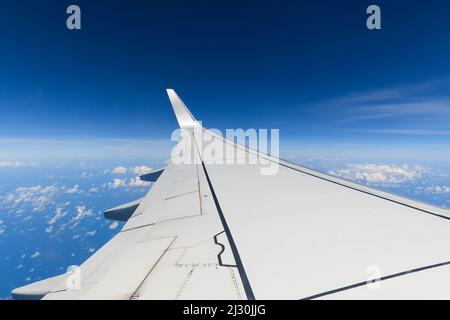 Blick auf den Flügel eines weißen Flugzeugs während eines Fluges an einem schönen blauen Himmel Tag mit einigen weißen Wolken, die hoch fliegen. Stockfoto