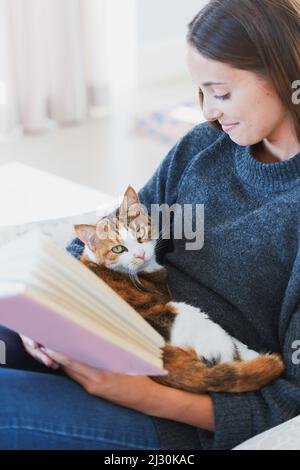 Glück ist eine flauschige Katze und ein tolles Buch. Aufnahme einer jungen Frau, die zu Hause ein Buch mit einer Katze auf dem Schoß liest. Stockfoto