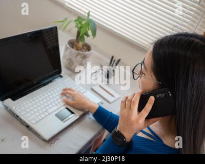 Asiatisches Mädchen mit Brille, Kopfhörer und Pflanzen, die von zu Hause aus mit einem Laptop und Handy arbeiten. Stockfoto