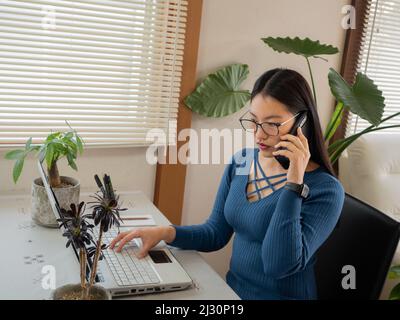 Asiatisches Mädchen mit Brille, Kopfhörer und Pflanzen, die von zu Hause aus mit einem Laptop und Handy arbeiten. Stockfoto