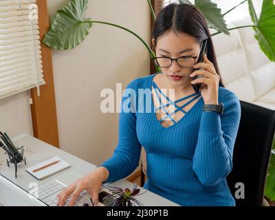 Asiatisches Mädchen mit Brille, Kopfhörer und Pflanzen, die von zu Hause aus mit einem Laptop und Handy arbeiten. Stockfoto