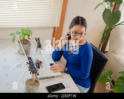 Asiatisches Mädchen mit Brille, Kopfhörer und Pflanzen, die von zu Hause aus mit einem Laptop und Handy arbeiten. Stockfoto