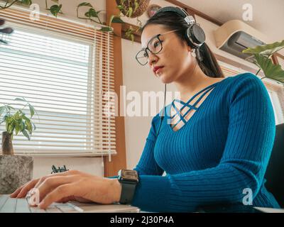 Asiatisches Mädchen mit Brille, Kopfhörer und Pflanzen, die von zu Hause aus mit einem Laptop und Handy arbeiten. Stockfoto