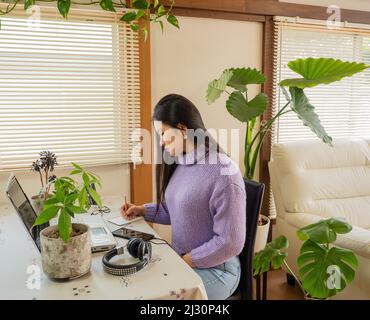 Asiatisches Mädchen mit Brille, Kopfhörer und Pflanzen, die von zu Hause aus mit einem Laptop und Handy arbeiten. Stockfoto