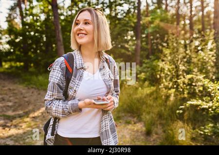 Junge Frau navigieren mit dem Mobiltelefon, während sie während der Sommerwanderung im Wald spazieren gehen Stockfoto
