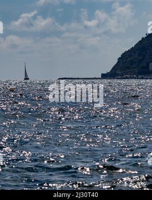 Ruhige Meereslandschaft mit einem einsamen Segelboot auf glitzerndem Wasser, mit einer zerklüfteten Küste im Hintergrund unter einem klaren blauen Himmel Stockfoto