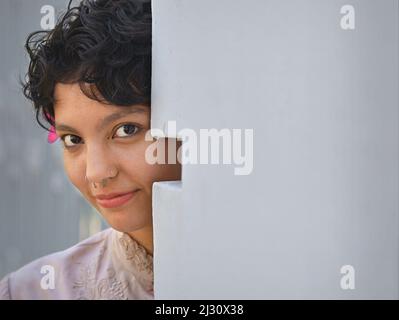 Schüchterne, hübsche junge Latina-Frau mit schönen großen braunen Augen und kurzen lockigen Haaren sieht vorsichtig um eine Ecke zum Betrachter hin. Stockfoto