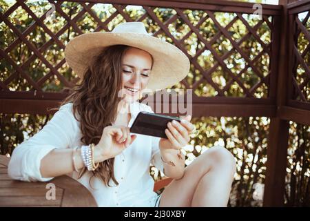 Überrascht moderne Hausfrau mittleren Alters in weißem Hemd mit Hut über Smartphone-App auf der Terrasse. Stockfoto