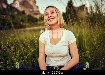 Porträt einer glücklichen, unbeschwerten jungen Frau, die auf der Wiese sitzt und den Abend in der Natur genießt Stockfoto