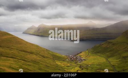 Blick auf ein einsames Dorf in einer weiten Landschaft. Funnings, Eysturoy, Färöer. Dunkle Wolken, Lichtblicke Stockfoto