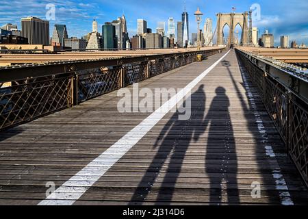 Die Schatten von zwei Freunden oder Liebhabern, die sich die Hände halten, die im Winter über die Brooklyn Bridge mit der Skyline von Lower Manhattan im Hintergrund laufen Stockfoto
