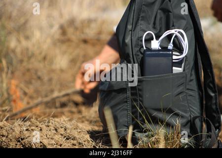 Smartphone mit schwarzer Powerbank im Rucksack aufgeladen. Eine Frau im Hintergrund zündet ein Lagerfeuer in der Natur an. Stockfoto