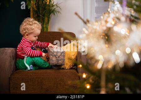 Am Weihnachtstag sitzt ein Kind auf einem Stuhl und packt ein Geschenk aus. Stockfoto