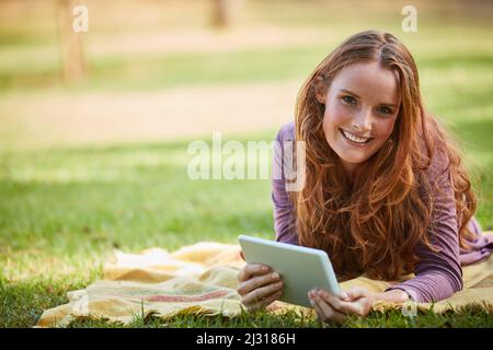 Ich kann mir keinen besseren Weg vorstellen, den Tag zu verbringen. Aufnahme einer jungen Frau, die ihr Tablet benutzt, während sie sich im Park entspannt. Stockfoto