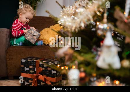 Am Weihnachtstag sitzt ein Kind auf einem Stuhl und packt ein Geschenk aus. Stockfoto