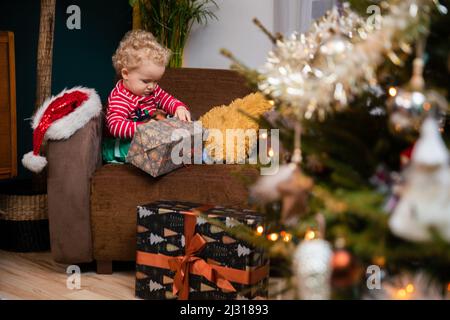 Am Weihnachtstag sitzt ein Kind auf einem Stuhl und packt ein Geschenk aus. Stockfoto