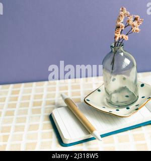 Notizblock und Stift, trockene Blume auf dem Schreibtisch. Lila Wandhintergrund. Arbeitsbereich Stockfoto