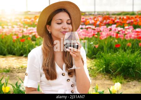 Schöne Frau riecht kostbaren Rotwein mit geschlossenen Augen im Garten mit blühenden Blumen auf dem Hintergrund Stockfoto