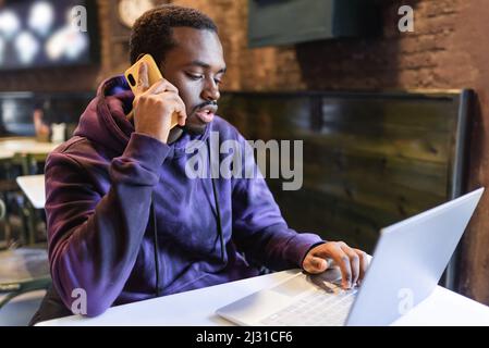 Junge afroamerikanische männliche Freiberuflerin, die am Handy spricht, während sie Remote-Arbeit auf Netbook im Café macht Stockfoto