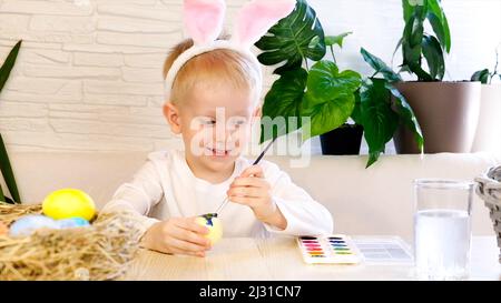 Ein kleiner Junge in Kaninchenohren malt begeistert Ostereier mit Farbe. Religiöse Feiertage Konzept, do it yourself, Familie Freizeit, osterei Stockfoto