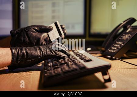Ein Mann in schwarzen Handschuhen mit einer Plastikkarte in den Händen an einer Computertastatur in einem dunklen Nachtbüro, Nahaufnahme Stockfoto