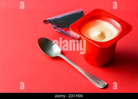 Cremig weißer, geöffneter Joghurt-Kunststofftopf. Isoliert auf rot mit selektivem Fokus und Kopierbereich Stockfoto