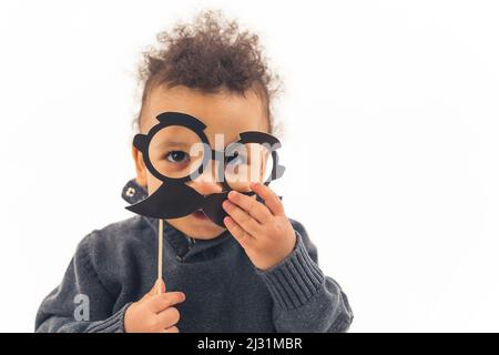 Schöne gewellte kleinen Jungen hält eine schwarze Maske aus Schnurrbart und Brille Blick in die Kamera Medium closeup Studio shot unendlich weiß . Hochwertige Fotos Stockfoto