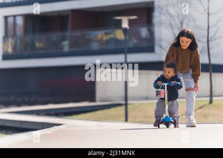 Schwarzhaarige dünne Frau, die einem kleinen afroamerikanischen Jungen mit lockigen Haaren folgt, die auf dem Roller stehen und seitwärts voller Schuss im urbanen Hintergrund aussehen. Hochwertige Fotos Stockfoto