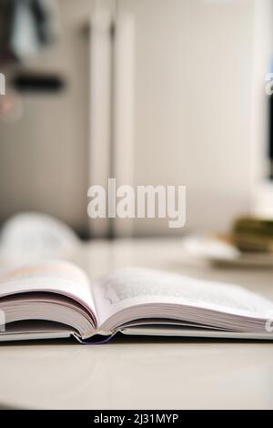 Offenes Hardcover-Buch mit weißen Seiten liegt auf Kunststeintisch in der Küche, Kühlschrank auf dem Hintergrund. Buchrücken. Selektiver Fokus. Vertikale Textabschnitte Stockfoto