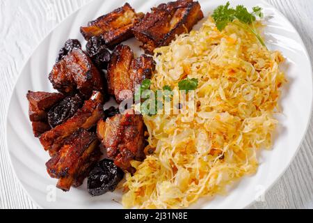 Geschmortes Sauerkraut mit Schweinefleisch und Pflaumen auf weißem Teller auf Holztisch, horizontale Ansicht von oben, Nahaufnahme Stockfoto