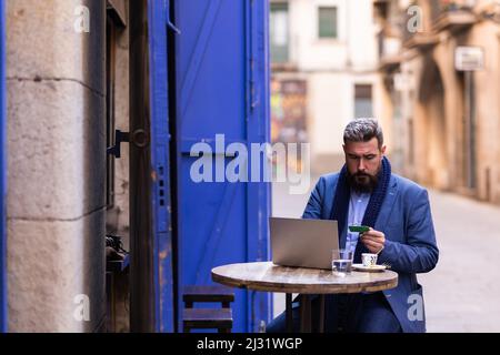 Mann, der draußen im Café sitzt und seinen Laptop und seine Kreditkarte benutzt Stockfoto