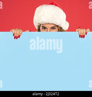 weihnachten, Weihnachten, Menschen, Werbung, Verkaufskonzept - glückliche Frau im weihnachtsmann-Helferhut, die hinter einem leeren blauen Brett auf einem roten Hintergrund guckt Stockfoto