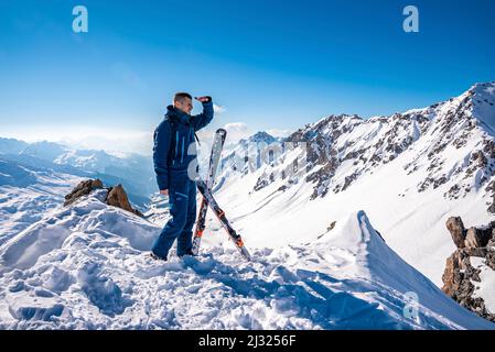 Junger Mann, der an sonnigen Tagen auf einem verschneiten Berg neben Skiern steht Stockfoto