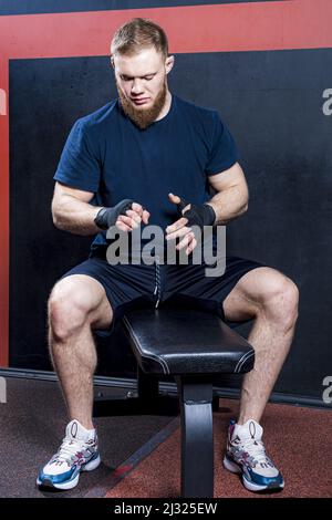 Der junge bärtige Kämpfer für gemischte Kampfkünste sitzt auf einer Sportbank und untersucht seine Hände, die in schützende elastische Binden eingewickelt sind. Stockfoto