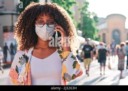 Junge schwarze Frau mit lockigen Haaren, die auf der Straße ein Mobiltelefon mit Gesichtsmaske verwendet Stockfoto