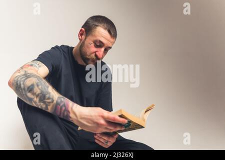 Hübscher bärtiger Kaukasischer tätowierte Mann, der auf dem Boden sitzt und die Heilige Bibel liest - Faith Konzept isoliert auf hellem Hintergrund mittlere Ganzaufnahme Studioaufnahme. Hochwertige Fotos Stockfoto