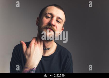 Charmanter europäischer Mann mit Tattoos Bart und Schnurrbart berühren Hals mit einer Hand und schauen zufrieden Medium Nahaufnahme isoliert auf gradienten grauen Hintergrund Studioaufnahme. Hochwertige Fotos Stockfoto