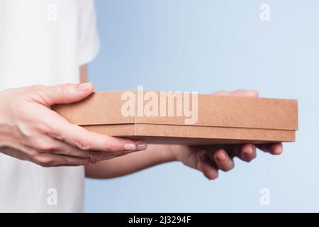 Frau Hände halten braunen ökologischen Verpackungskarton aus natürlicher Wellpappe. Mockup-Versandkarton auf blauem Hintergrund. Verpackung Stockfoto