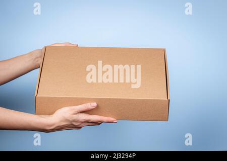 Frau Hände halten braunen ökologischen Verpackungskarton aus natürlicher Wellpappe. Mockup-Versandkarton auf blauem Hintergrund. Verpackung Stockfoto