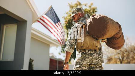 Rückansicht eines patriotischen jungen Soldaten, der mit seinem Gepäck auf sein Haus zuläuft. Mutiger amerikanischer Militärangehöriger, der nach seinem Einsatz in der wieder nach Hause kommt Stockfoto