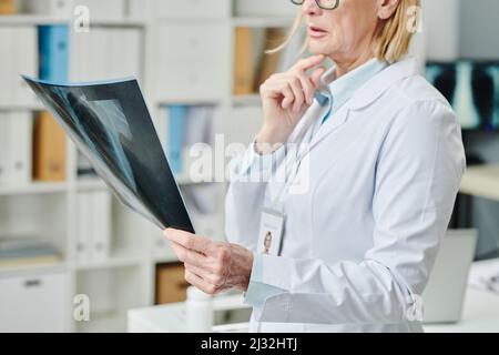 Moderne reife Ärztin in Weißkohlragen, die das Röntgenbild der Lunge vor sich hält und an die Diagnose denkt Stockfoto