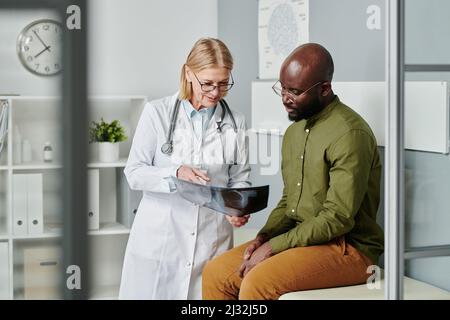 Selbstbewusster reifer Arzt in Weißkohlensäurebild, der einen jungen schwarzen Mann mit einer Röntgenaufnahme seiner Lunge zeigt und ihm die Diagnose in Kliniken erklärt Stockfoto