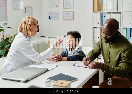 Reife Kinderärztin misst die Körpertemperatur eines kleinen Patienten mit einem elektronischen Thermometer, während ein junger Mann die medizinische Form ausfüllt Stockfoto