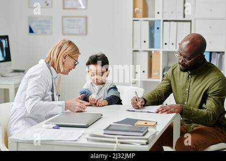 Junger afroamerikanischer Mann, der medizinische Formulare ausfüllt, während er am Schreibtisch vor seinem kleinen Sohn und Arzt sitzt und Pulsoximeter verwendet Stockfoto